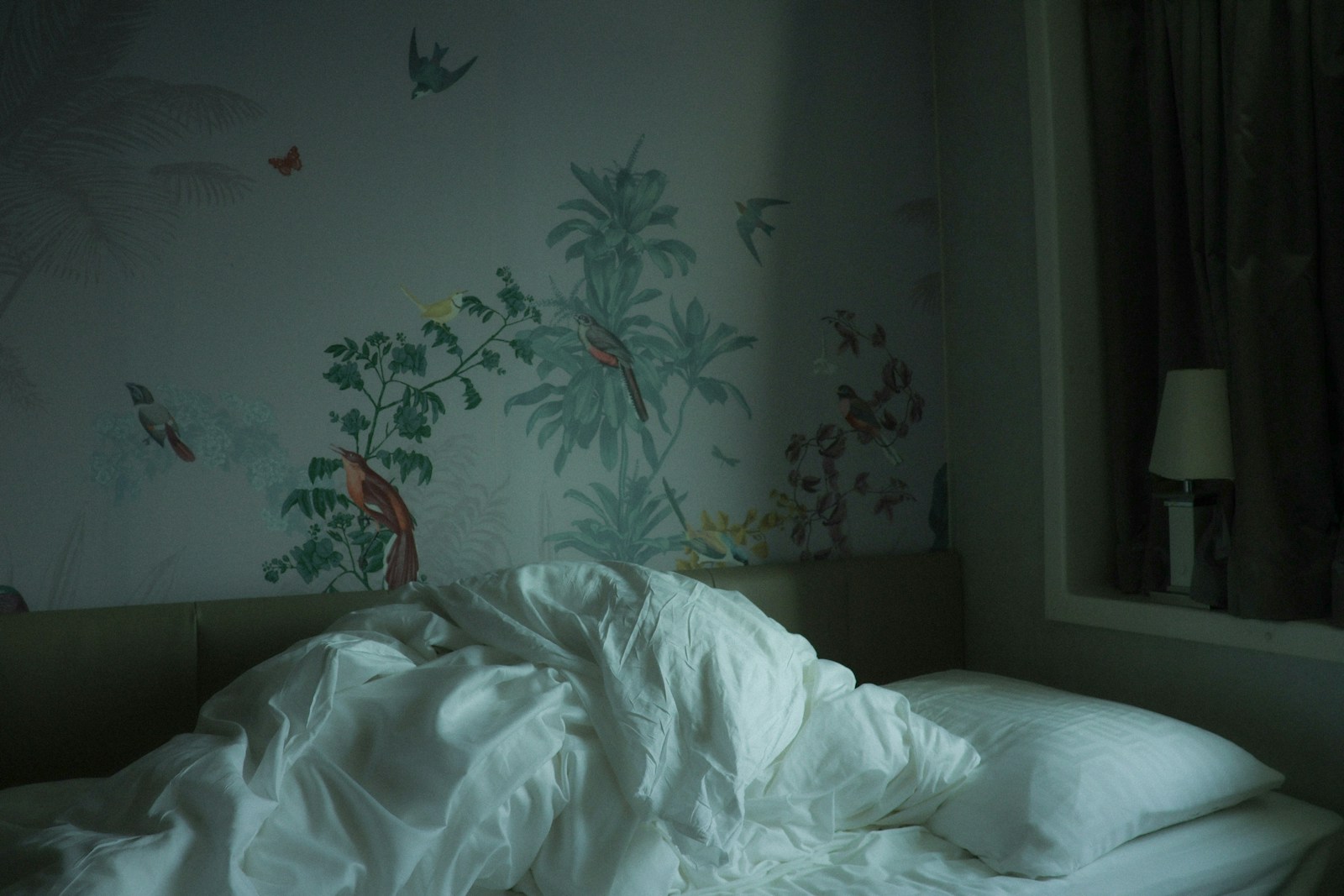 An unmade bed with white bedding in a dim bedroom beside a curtained window.